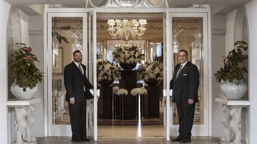 Doormen open front entrance doors to lobby with white flower arrangements on pedestals