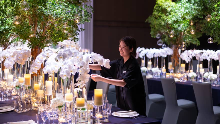 A wedding staff member preparing a table with elegant glassware and candlelights, arranging the white floral centrepiece to create a fresh and inviting setting.