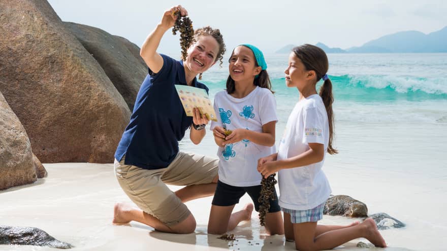 Woman kneels on white sand beach beside boulder, holds up seaweed to show two girls