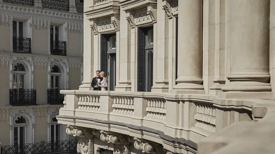A couple in formal attire stands on a grand balcony of an elegant building, sharing a moment while holding champagne glasses, with ornate architecture in the background.