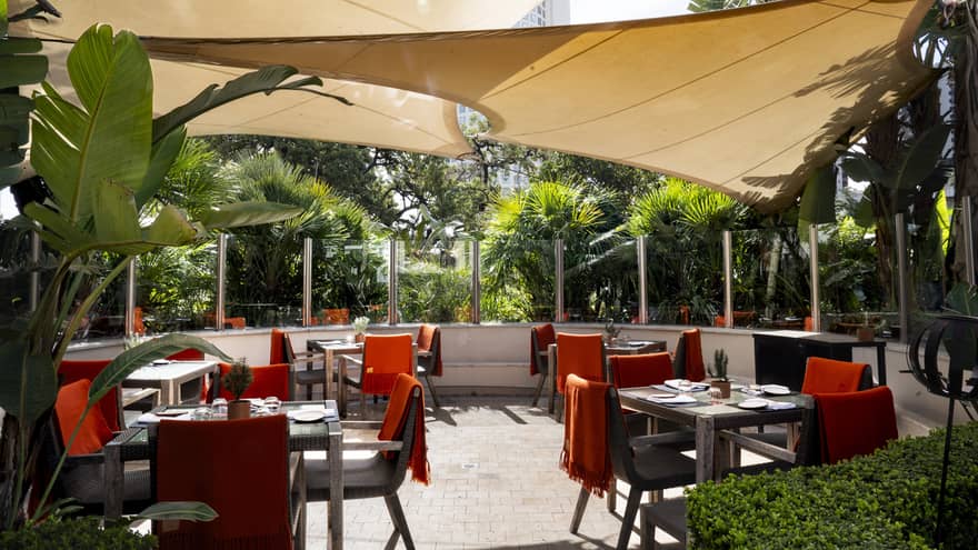 Outdoor dining patio with black-and-red chairs and square tables with three beige sun shades hanging overhead and lush greenery surrouding the patio