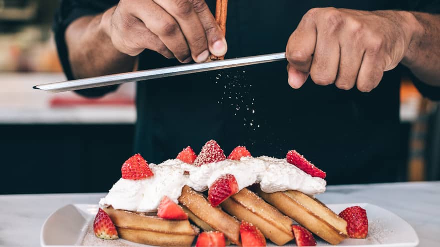 Close-up of waffles topped with whipped cream and strawberries, chef shaving cinnamon stick