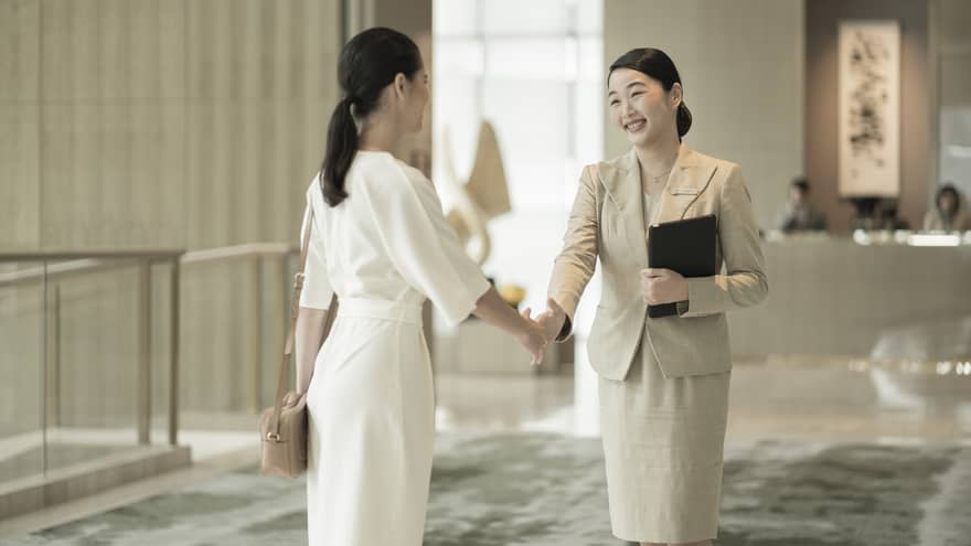 Two women shake hands in a lobby with grey carpeting, in front of a welcome desk.