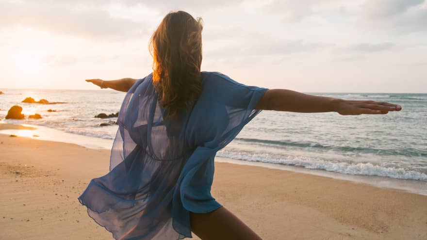 Woman wearing sheer beach cover kneels with arms outstretched in yoga pose on sandy beach by water
