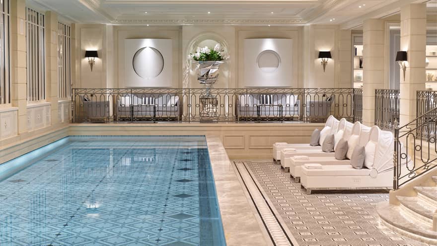 Long blue indoor swimming pool with large tile deck, steps, white lounge chairs