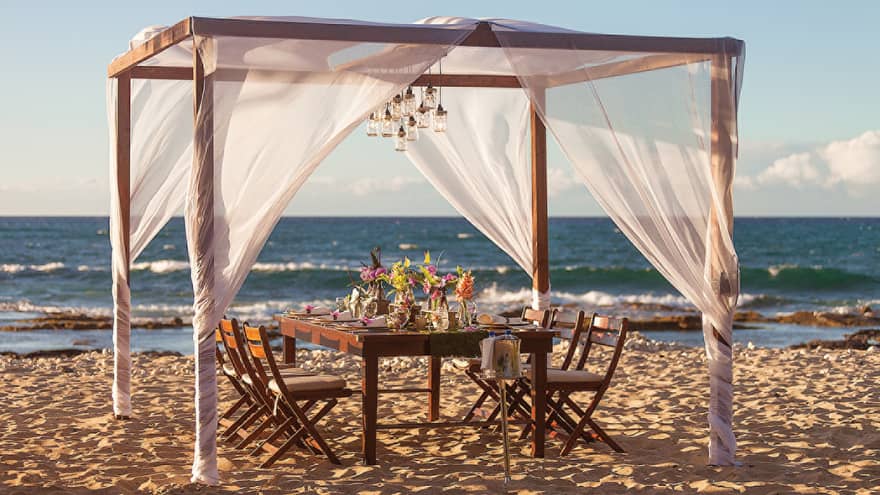 Wood dining table with fresh flowers under sheer white canopy, lanterns on beach