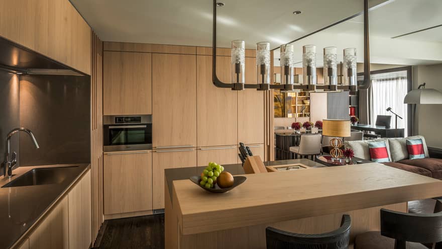 Modern kitchen area with light wood cabinetry, a built-in oven, sink and a central island with a fruit bowl, leading into a cozy Four Seasons living space