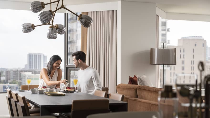 Smiling couple enjoys breakfast in their guest room, views of downtown Houston behind them 