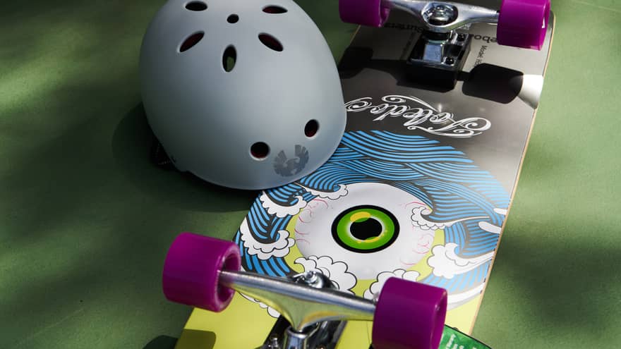 Skateboard with purple wheels and a yellow-and-blue ocean design with a large eye in the middle sits on a green surface with a grey helmet next to it
