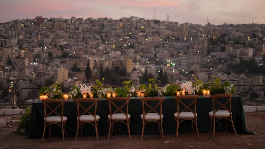 Rectangular table with dark linens, florals and candles lit in front of each place setting, and a row of six wooden chairs with white seats all in a row facing a city below lit up beneath a pink-and-blue sky at dusk