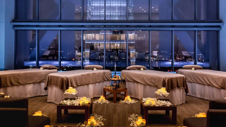 Four message beds in a dimly lit room with yellow flowers laid around the floor, large windows show a city in the background.