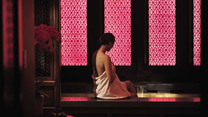 Woman wearing white towel sits on edge of dark spa tub by wall with pink lights