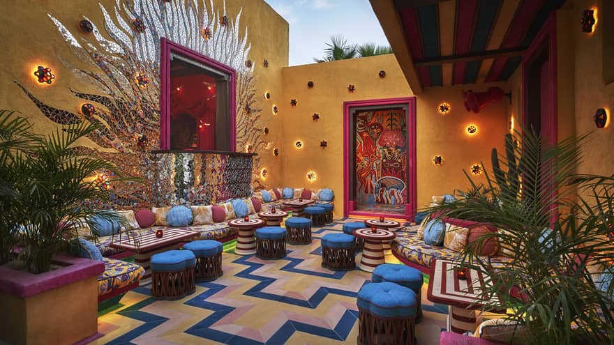 A colourful outdoor seating area with bright blue cushioned stools, colourful lights and plants.