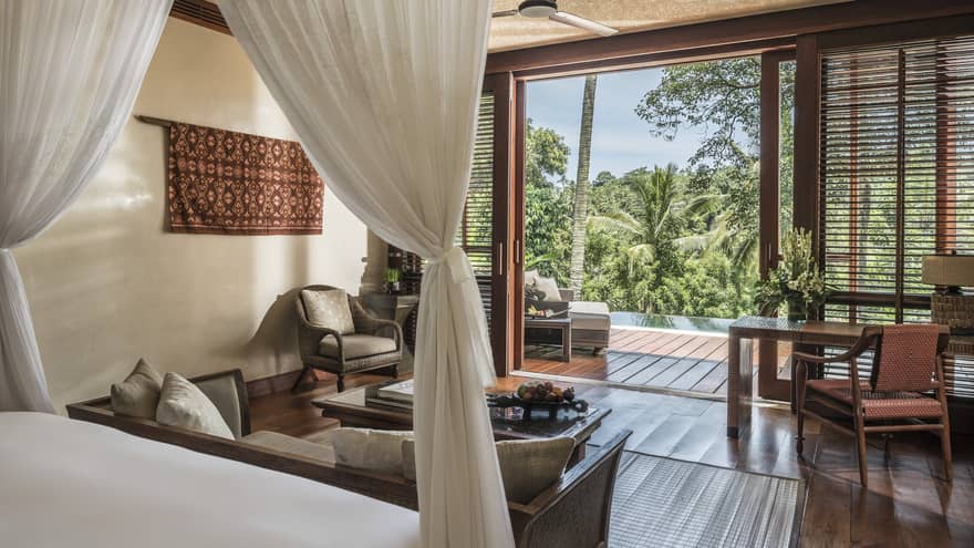 Villa bedroom with canopy bed, hardwood floors, desk and desk chair, arm chair, sliding door to pool and balcony