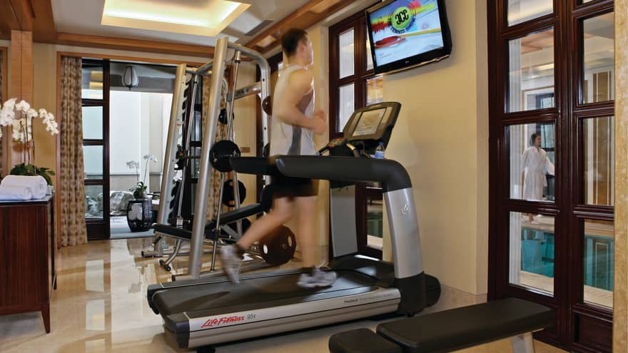 Man running on treadmill in Fitness Centre, woman in robe walking by pool in background