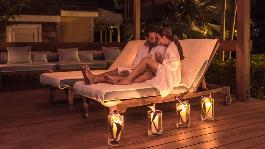 Man and woman in bathrobes relax on lounge chairs, glowing candles on deck at night