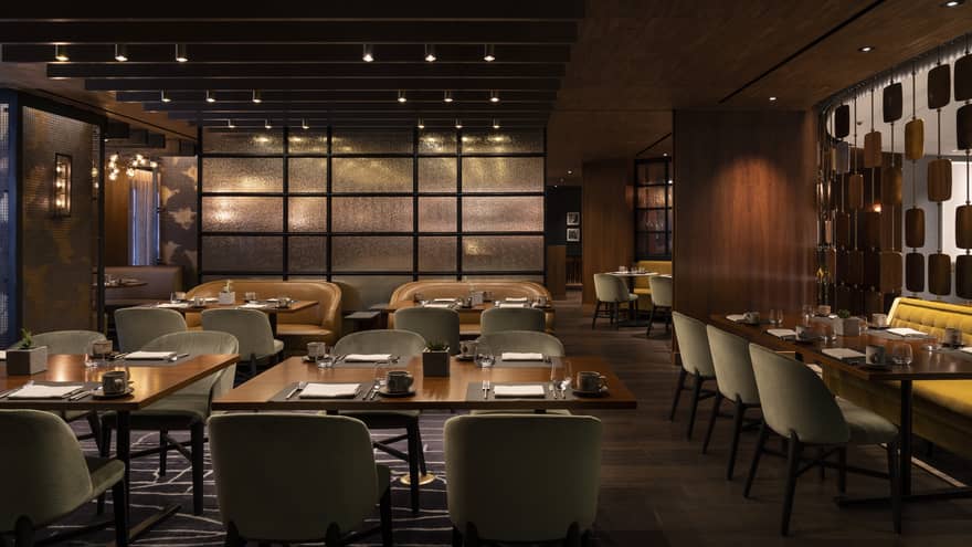 The interior of a dimly lit restaurant with square tables, grey chairs and wood walls.