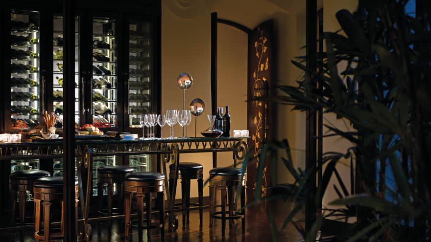 Wine bottle, glasses, cheese platter on table in front of wine cellar in dark restaurant