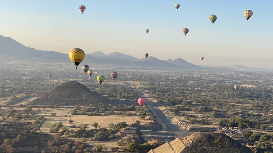 An aerial view of colourful hot-air balloons floating over pyramids and trees, with mountains and a city in the background.