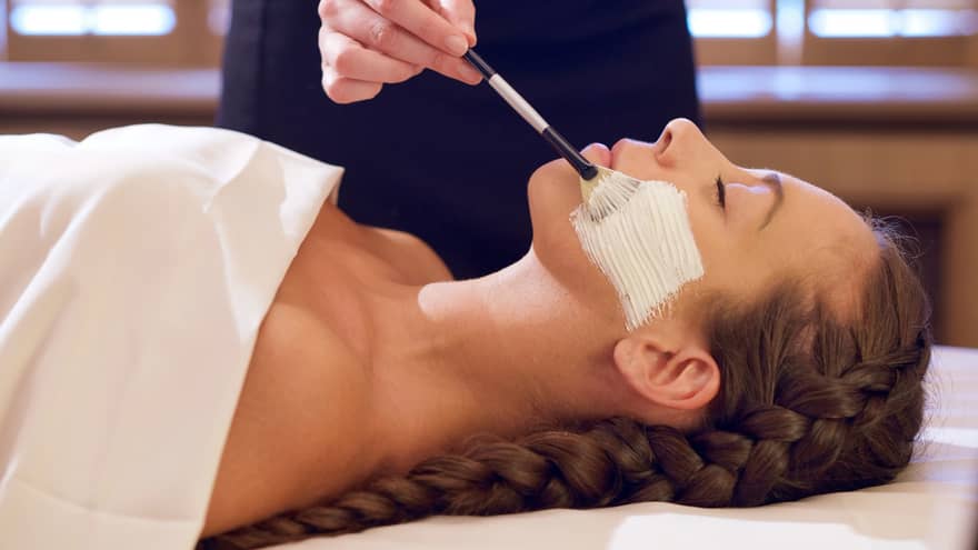 Woman with eyes closed lies on back on spa table as woman with paintbrush applies Sodashi-based facial cream