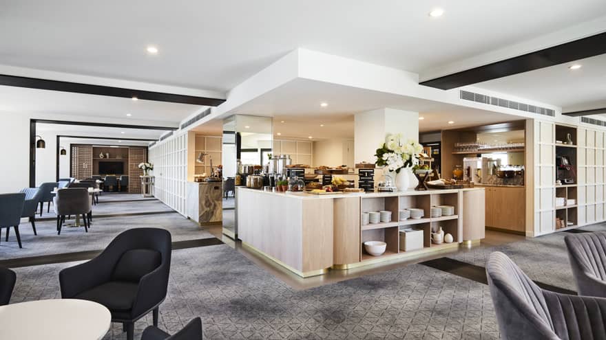 A modern hotel lounge with plush grey chairs and carpet, and a well-equipped kitchen set up with buffet foods and coffees.