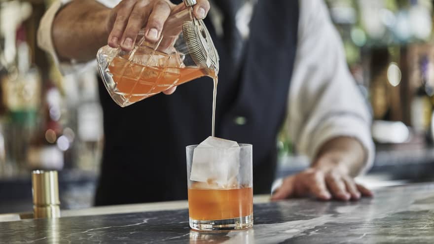 Close-up of bartender pouring orange cocktail from shaker into crystal glass with large ice cube
