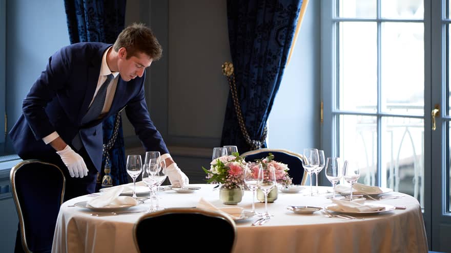 Hotel staff wearing white gloves places dish on small round dining table