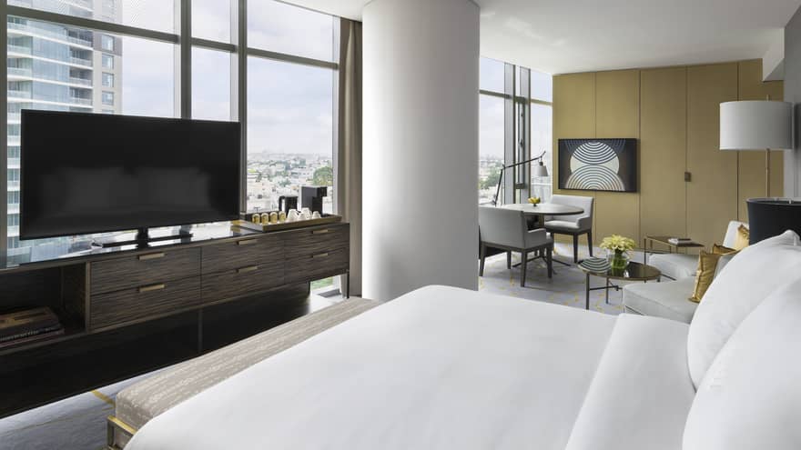 Premier Room with white bed facing large flat-screen TV on console, and floor-to-ceiling windows with city views