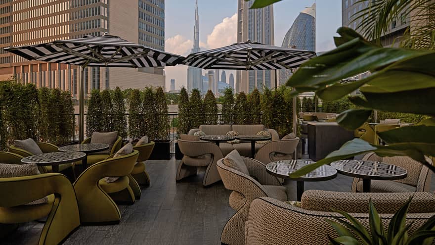 Outdoor restaurant patio filled with small bistro tables, beige upholstered chairs, black-and-white striped umbrellas and a hedge of bushes lining the ledge
