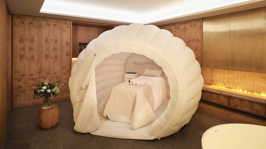 A spa treatment table with white draping and pillow within a near-spherical white tent, all in a spacious, softly lit treatment room with decor in warm earthy tones