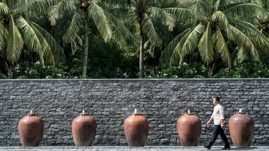 Man walks by palm trees, tall stone fence lined with large clay pot fountains