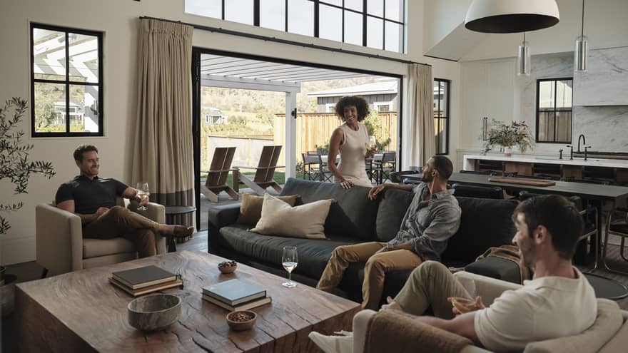 Friends gather around a table in a living room, seated on a couch and club chairs, with a view of the villa's outdoor area.