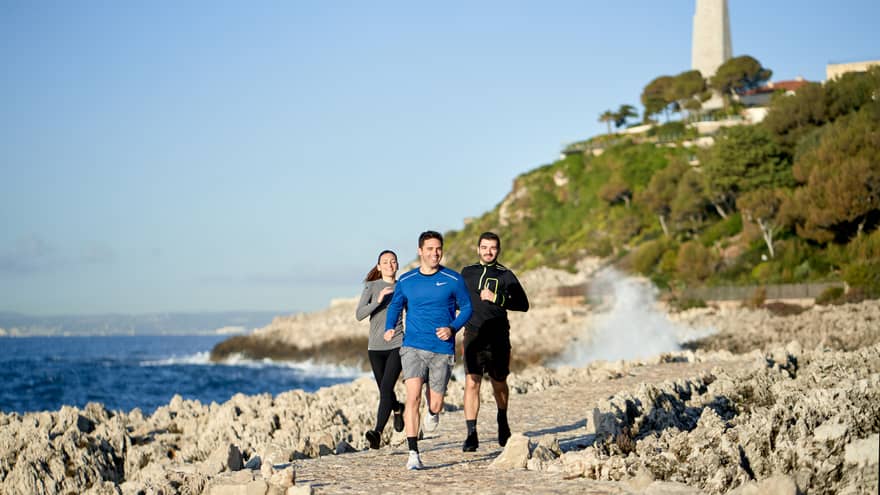 Three people jogging, smiling, on rocky trail beside the sea, lighthouse in backdrop