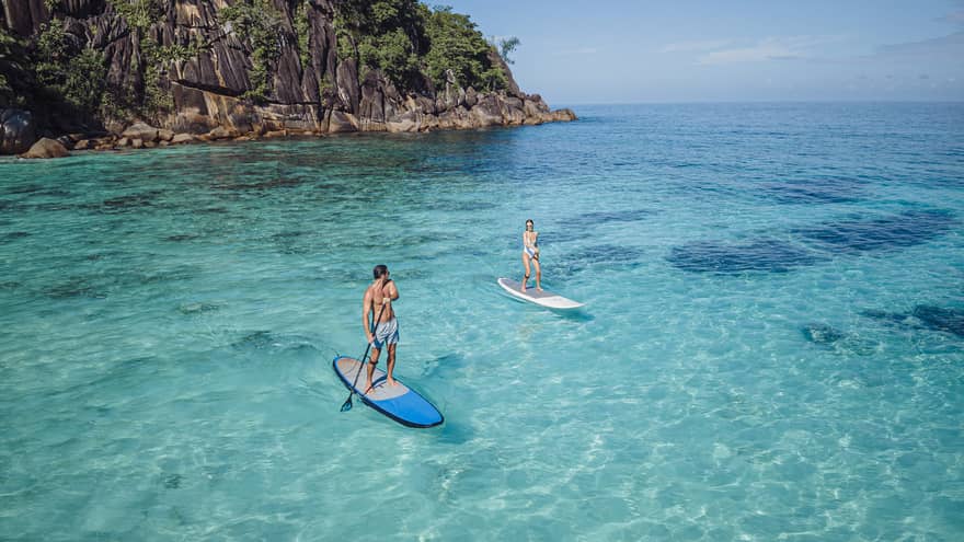 Two people stand-up paddleboarding on clear blue water with rocky cliff in back drop