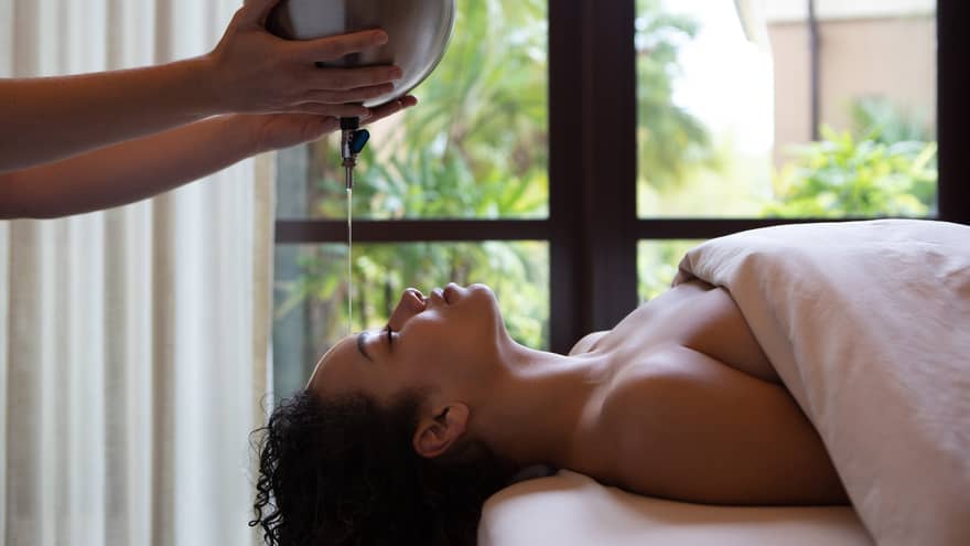 A person lies down with eyes closed, receiving a massage. Warm oil gently pours on their forehead as natural light streams through the curtains, creating serenity.