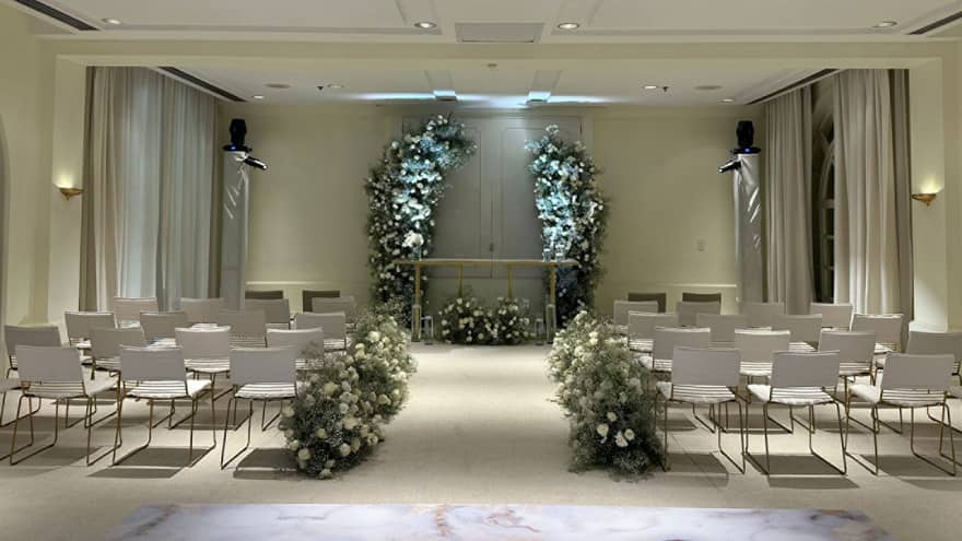 A spacious, softly lit indoor wedding space with rows of white chairs and floral decor accenting serene neutral interior finishes.