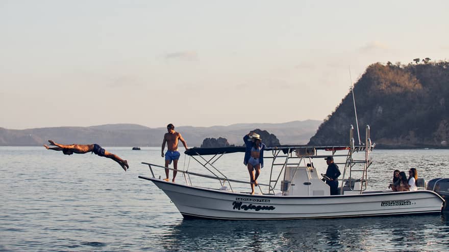 Side view of a guest diving gracefully into the water while others watch from a boat's deck, distant landmasses beyond.