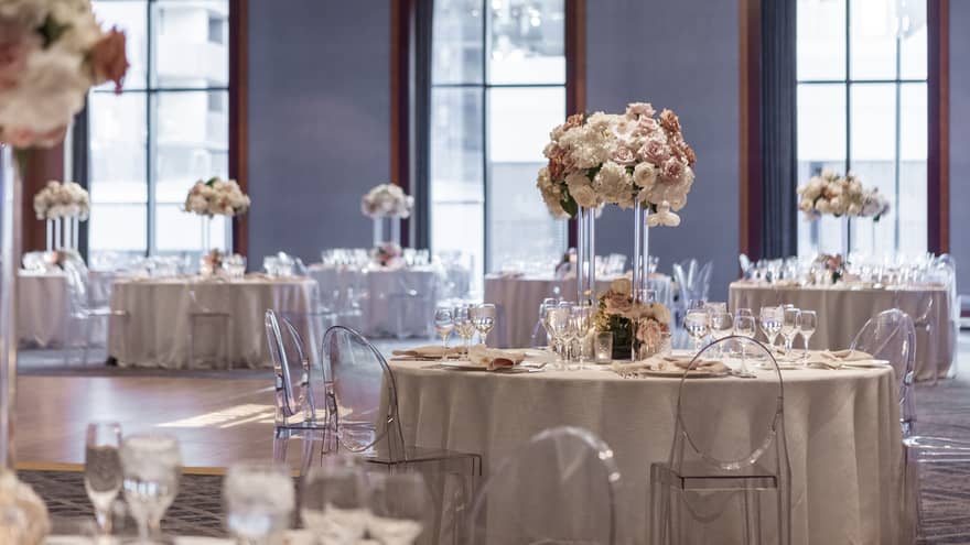A ballroom with round tables with grey covers, flower centerpieces, and clear chairs.