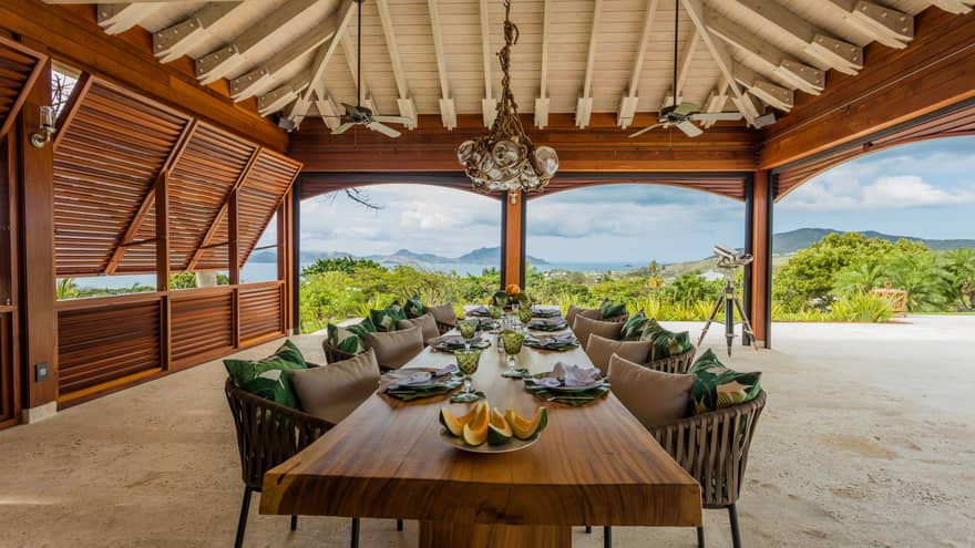Melon slices on long rustic wood dining table, chairs and pillows under covered beach patio