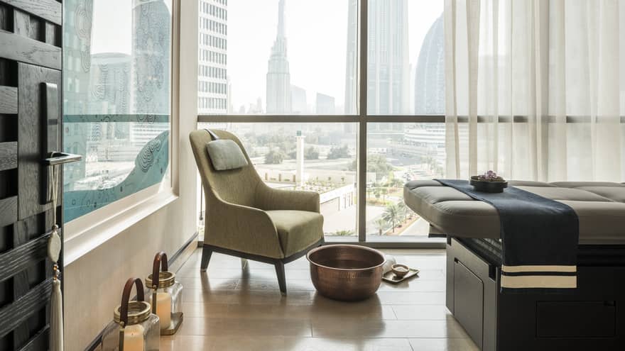 Armchair in hotel room corner by sunny window, large brass bowl foot bath, rolled white towel on floor