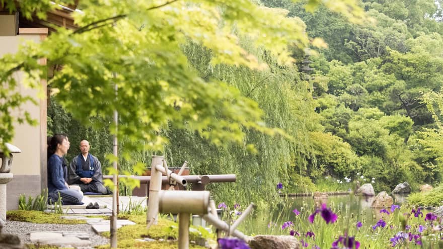 Two people sit cross-legged, in meditation, overlooking a pond surrounded by green vegetation in Kyoto