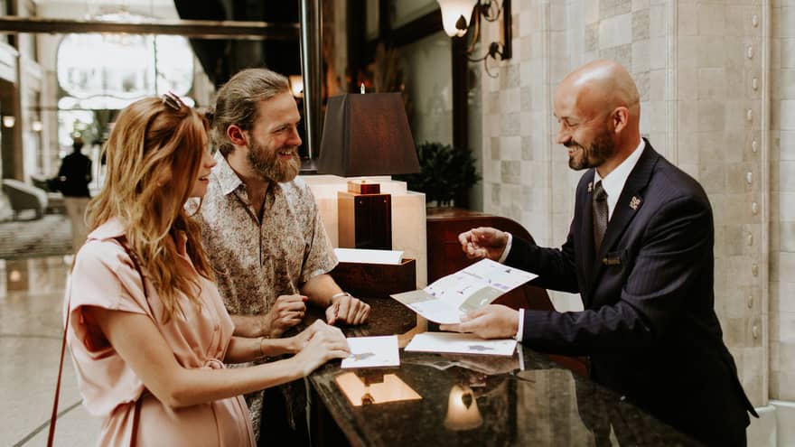 Couple at a hotel reception desk speaking with a Concierge, who is showing them a brochure.