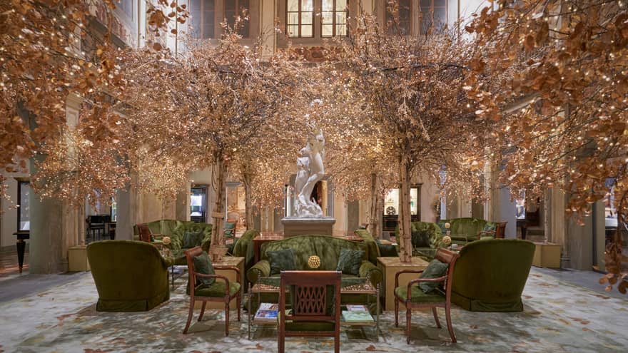 A large two-storey lobby is decorated with green velvet couches and chairs, golden-leafed trees, and a white marble statue.