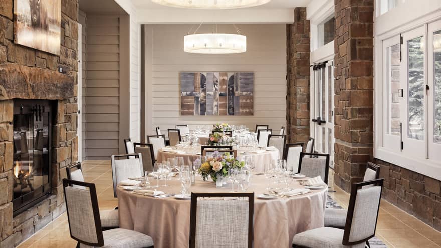A row of round tables set for a wedding, featuring floral centrepieces, cushioned chairs, a stone fireplace and large windows 