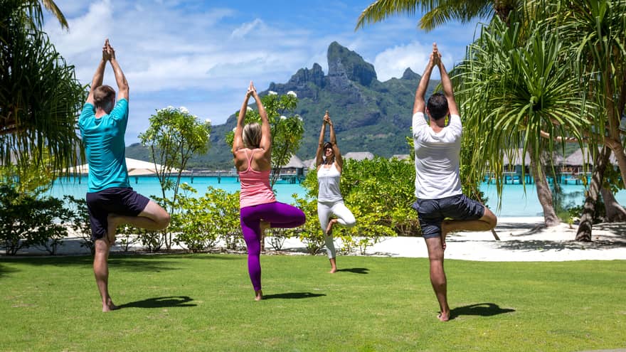 Three guests and an instructor practice yoga on the lawn, the lagoon and mountains visible behind them