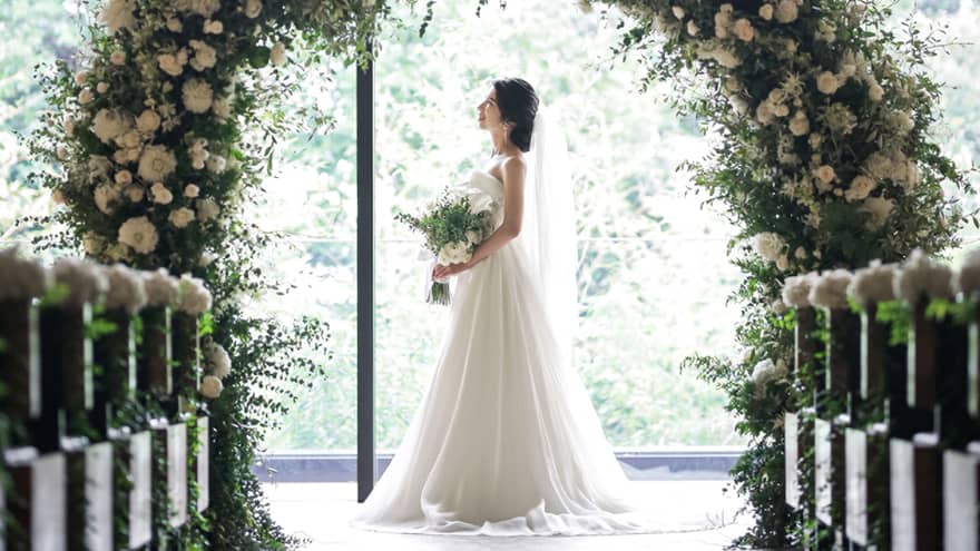 Bride stands under round wedding altar covered in white flowers by sunny window