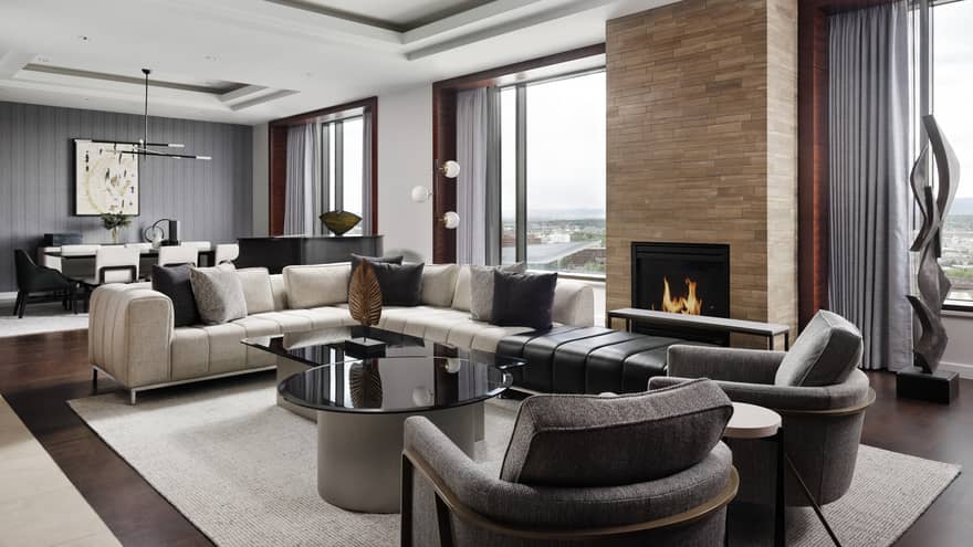 A lounge seating area with a fire place, a white sofa, two grey chairs, and a black table between them; there are also large windows.