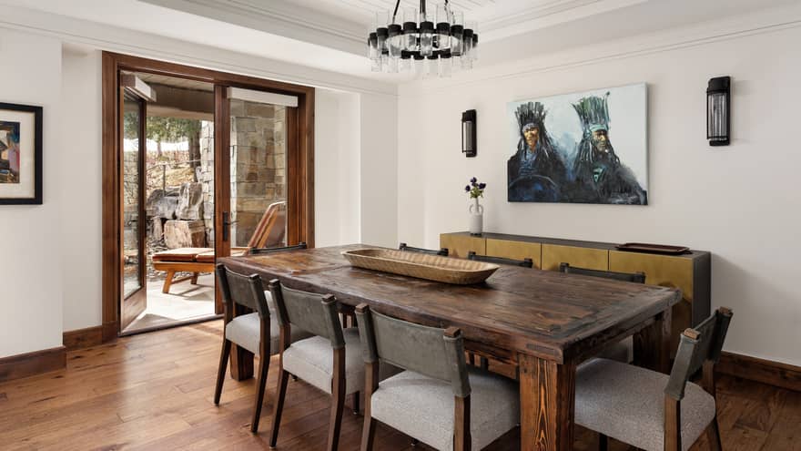 Dining room with rustic wooden table, eight chairs, large painting, glass chandelier and open doors to stone patio