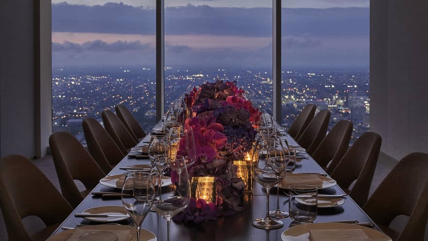 A long dining table with red flowers in the center, a sun set can be seen above a city through a large window.