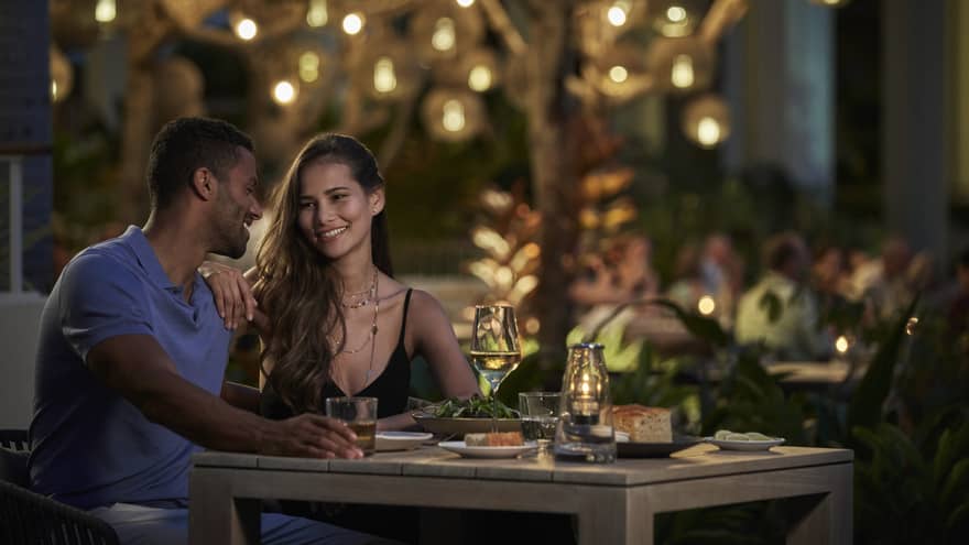 A couple sits together at a dining table beneath a tree lit with twinkling string lights